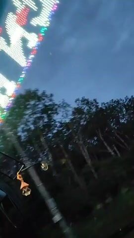 This is a first-person perspective video of a bus ride in Bờ Y, Vietnam, during dusk. The camera is positioned inside the bus, looking forward through the windshield. A string of colorful, bright LED lights is visible along the top edge of the windshield, suggesting the bus is decorated or part of a festive occasion. Outside the bus, the landscape is dominated by lush green trees and vegetation, characteristic of a tropical environment. The sky is a deep blue, indicating the transition from day to night, with a few faint clouds.

The bus appears to be traveling on a paved road, which is visible in the lower portion of the frame. The motion of the bus causes the scenery to blur, creating a sense of movement and speed. A dangling decorative item, possibly an air freshener or charm, hangs from the rearview mirror, swaying gently with the bus's movement. There are no people clearly visible within the frame other than the implied presence of the bus driver. The overall mood is one of travel and observation, with the colorful lights hinting at a lively atmosphere. The location cues, such as the dense vegetation and the appearance of the road, are consistent with a rural or semi-rural setting in Vietnam.