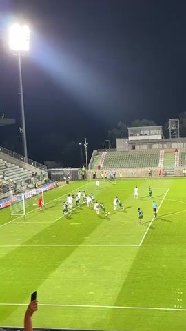 A soccer game is being played at night under bright stadium lights. Two teams, one in white uniforms and the other in green uniforms, are on the field. The white team is on the offensive and has possession of the ball near the goal. Several players are gathered around the goal mouth, jostling for position. A player in white appears to be attempting a shot or a pass. A player in green, presumably the goalkeeper, is positioned in the goal. Spectators are visible in the stands on the left and right sides of the stadium, with some holding up phones. The field is well-lit and appears to be artificial turf.