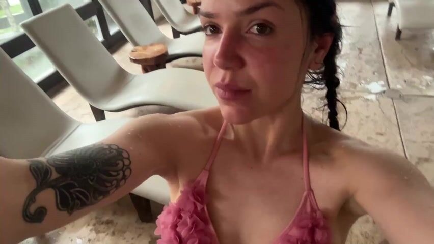 A woman with wet hair and a tattoo on her left upper arm, wearing a pink floral bikini top, is recording a video of herself in an indoor pool area. There are rows of empty white lounge chairs and a small pool with rocks in the background. The floor is wet. She looks into the camera and touches her hair.