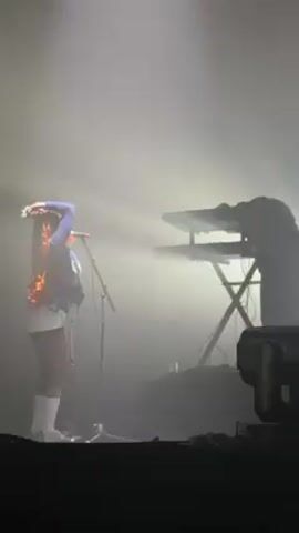 On a hazy stage, a woman stands with her back to the camera, facing stage right. She has long, dark braids with orange highlights and wears a blue long-sleeved shirt, a vest, white shorts, and white socks. She holds her hands above her head, fixing her hair, and a microphone stands in front of her. To the right, a man in dark clothing stands behind a keyboard, his face obscured by the lighting. A spotlight shines on the singer, creating a silhouette of the keyboardist. Stage equipment is visible in the foreground.