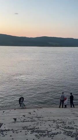 A wide river flows calmly under a pale pink and blue sky. A concrete embankment slopes down to the water. On the embankment, four people are visible. On the left, a person wearing a patterned garment is bent over, possibly tending to something on the ground. Further to the right, three figures stand together, two adults and one child. The adult on the left is wearing a light-colored shirt and dark pants. The child is wearing a white coat. The adult on the right is wearing a dark jacket and dark pants. The figures are positioned facing the river, with the two adults and child looking out at the water. The environment appears to be outdoors, possibly at dusk or dawn, with a gentle breeze creating ripples on the water's surface. The embankment is scattered with small rocks and debris.