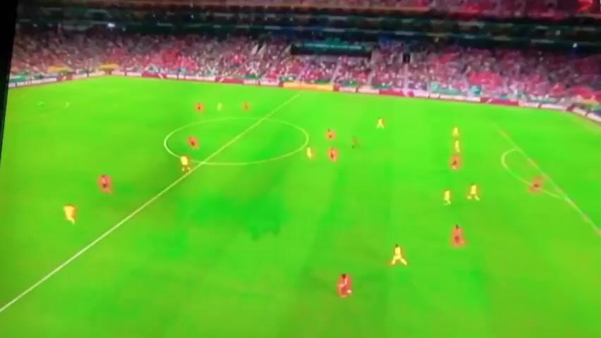 A soccer game is being played on a bright green field. There are multiple players wearing red and yellow jerseys. The players are spread out across the field, with some clustered around the center circle and others near the goal. A goalkeeper in a white jersey is positioned in front of the goal. The stands are filled with spectators. The Qatar Airways logo is visible on a banner behind the goal.