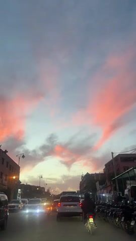 The video shows a traffic scene at dusk in what appears to be a city in Morocco. The sky is filled with pink and grey clouds, indicating either sunrise or sunset. The view is from behind the windshield of a vehicle, with several cars and motorcycles visible on the street. Buildings line both sides of the road, with street lights illuminating the scene. A person on a motorcycle is in the foreground, moving along with the traffic. The overall atmosphere suggests a typical evening commute in an urban environment.
