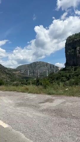 The video shows a long, white concrete bridge stretching across a valley, connecting two mountains. The bridge is supported by tall pillars. The valley is covered in green trees and vegetation. The sky is blue with white, puffy clouds. The camera is moving slowly along a dirt road, with tall grasses and shrubs in the foreground. The wind is blowing strongly.