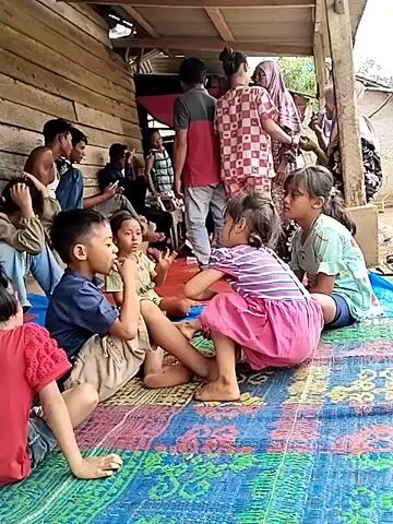 A group of people, primarily children, are gathered outdoors under a wooden structure. The ground is covered with a colorful mat, and a blue tarp is visible. Several children are sitting on the mat, interacting with each other. In the foreground, two children are engaged in play. A boy in a blue shirt and tan shorts is sitting on the left, and a girl in a pink and purple striped dress is on the right. They are touching each other's faces and appear to be smiling or laughing. Other children are seated nearby, observing or participating. In the background, several adults are standing and conversing. The setting appears to be a rural or informal environment, possibly a village or a makeshift shelter, with wooden walls and a corrugated metal roof. The lighting suggests it is daytime.