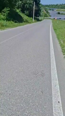 The video shows an upward-sloping paved road with a white line marking the right edge. The road is surrounded by green grass and trees. In the distance, there is a body of water, a bridge, and a mountain. The sky is blue with some clouds. The camera appears to be moving forward along the road, possibly from the perspective of a vehicle.