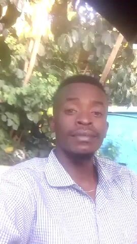 A man with dark skin and short black hair is facing the camera. He is wearing a light blue and white striped short-sleeved shirt. The background is blurry but appears to be outdoors with green foliage. He is speaking.