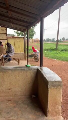 A person wearing a red hooded sweatshirt and gray pants is using a wheelbarrow. They are bent over the wheelbarrow, appearing to be placing something inside it. The wheelbarrow is on a dirt path. In the background, there are buildings, trees, and a cloudy sky. In the foreground, there is a concrete structure and a wooden bench.