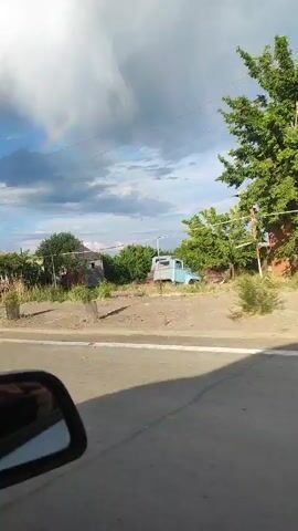 A car drives along a road. The driver's side mirror is visible in the bottom left corner. The road is asphalt with a white line down the middle. On the right side of the road, there are houses and buildings. Trees are scattered along the road. In the background, there are clouds in the sky, and a rainbow is visible on the left side. The car passes by a construction truck and a black car. A power pole and power lines are also visible.