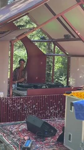 A person wearing sunglasses and a patterned shirt stands behind DJ equipment on a raised stage in a wooded area. Bubbles are floating in front of the stage. Speaker equipment and wires are visible on the floor in front of the stage.