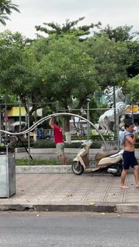A man wearing a red shirt and light-colored shorts is standing on a sidewalk and holding onto a curved metal structure. He appears to be pushing or adjusting the structure with his hands. Behind him, there is a scooter parked. The background shows trees, a white horse statue, and other people in the distance. The overall environment seems to be an outdoor public space, possibly a park or a plaza. The lighting suggests it is daytime.
