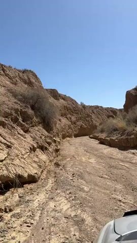 A first-person point-of-view shot from a vehicle shows it driving along a dry, eroded dirt track. The track is flanked by steep, rocky, light brown hills or cliffs on both sides, characteristic of an arid environment. Sparse, dry vegetation is visible on the hills. The sky is clear and bright blue, indicating sunny weather. The scene depicts a journey through a canyon-like landscape, likely in Ak Say, Kyrgyzstan. The front part of a silver or white vehicle is partially visible in the lower right corner of the frame. No text is visible.