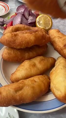 The video shows a close-up of golden-brown fried dough pastries arranged on a plate. In the background, a salad of red onion, tomato, and lettuce is visible, along with a lemon wedge and a knife. A spatula is used to lift one of the pastries from a pile, revealing its crispy texture. The pastries appear to be filled and are served with fresh vegetables and a lemon slice.