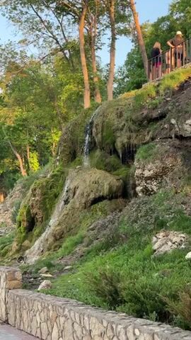 The video shows a small waterfall cascading down a moss-covered rock face. The waterfall is thin, with water trickling down the rocks. The rock face is covered in lush green moss and some vegetation. At the top of the rock face, there is a railing with people standing behind it. The background includes tall trees with green leaves and a clear blue sky. A stone wall lines the base of the rock face. The light suggests it is late afternoon or early evening.