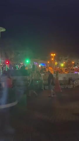 The video shows a night scene in what appears to be an outdoor entertainment area or promenade. There are several people present, some walking and others gathered. Lights, including green, purple, and orange, illuminate the area, creating a festive atmosphere. In the background, there seems to be a stage or raised area with people on it and a crowd gathered. There are trees and bushes, and the ground is paved with what looks like brickwork. Some streetlights are also visible. The overall impression is of a lively evening gathering.