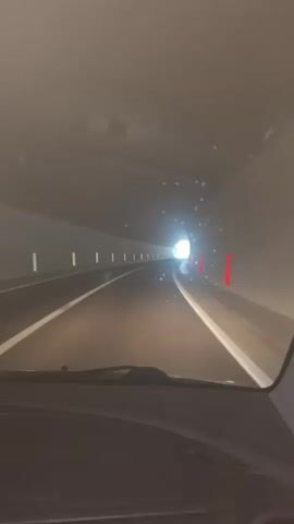 The video shows a first-person perspective of driving through a tunnel. The tunnel is dark and dimly lit, with bright lights visible at the end, suggesting the exit is approaching. The road has white lane markings. Orange reflective markers are visible on the right side of the tunnel, and white reflective markers are on the left. Small white specks are visible in the air throughout the tunnel. The interior of the car, including the dashboard and windshield wipers, is visible in the foreground. The time of day and weather are not explicitly stated but the dim lighting suggests it might be nighttime or heavily overcast. The location is identified as Strp, Montenegro. No text is visible in the video. The overall mood is one of progression and anticipation as the car moves towards the light at the tunnel's end.