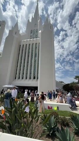 The media file captures an outdoor scene in front of a large, white, modern temple-like building, likely a church or place of worship. The architecture is striking with tall spires, sharp angles, and numerous vertical windows. The building appears to be under a bright, partly cloudy sky, suggesting it's daytime with pleasant weather.

A crowd of people, dressed in a variety of attire from casual to more formal, are gathered on the grounds in front of the building. Some are standing in groups, engaged in conversation, while others are walking or sitting. Children are also present, some in what appear to be special occasion dresses. The overall atmosphere seems to be one of a gathering or event, possibly a ceremony, celebration, or open house.

The foreground is filled with lush greenery, including large, spiky plants and tall, leafy foliage, indicating a well-maintained garden area. In the middle ground, the crowd is the central focus, with individuals interacting and milling about. Behind the crowd, glimpses of other buildings and palm trees can be seen, suggesting an urban or suburban setting in a warm climate. The presence of white tents or canopies suggests some sort of temporary setup for the event.

Notable details include the bright daylight illuminating the scene and casting shadows, and the distinct architectural style of the main building. The people's body language and interactions suggest a positive and perhaps celebratory mood. The setting, with its manicured landscaping and large gathering, points to a significant occasion.

There is some visible text on the building above the entrance which reads: "THE HOUSE OF THE LORD" and below it, "HOLINESS TO THE LORD". The location context provided is San Diego, United States of America, which aligns with the sunny weather and palm trees visible.