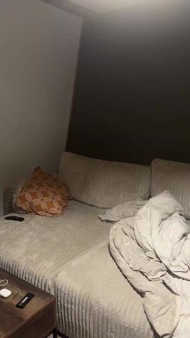 A cat jumps off a couch and runs towards a cat tree. The couch is beige and made of corduroy material. There is an orange patterned pillow on the couch, and a white blanket is partially folded. There is a brown side table to the left of the couch with a remote control and what looks like wireless earphones. The cat is orange with white markings. The floor is wood. The cat climbs onto the cat tree.