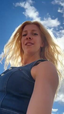 The video begins with a close-up shot of a blonde woman with her eyes closed, smiling brightly, against a backdrop of a bright, sunny sky with a few clouds. The sun is positioned in the upper right corner, creating a lens flare that obscures some details. She is wearing a denim outfit and a necklace with star-shaped pendants.

As the video progresses, the camera pans out to reveal she is standing in a field of dry, golden grass or crops under a blue sky with scattered clouds. She is smiling and her arms are slightly outstretched, as if embracing the surroundings.

In the latter half of the video, a white car with a red roof comes into view, positioned to her right. The woman continues to smile and her hair blows in the wind. The background appears to be a rural landscape with fields and trees visible in the distance. The sun remains a strong presence, casting a bright light on the scene. The camera angle is low, looking up at the woman.