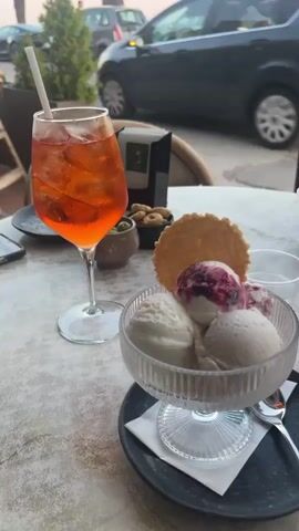The video shows a table on a patio, with ice cream, a drink, snacks and a chocolate milkshake, and people seated at the table. Cars are parked on the street nearby. Palm trees line the edge of the beach in the background. The cars are primarily a dark grey SUV, a white SUV, and a red SUV. The sky is pink and blue, indicating either sunset or sunrise.