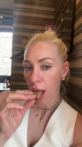 A woman with blonde hair pulled into a bun is eating a small piece of food. She is wearing a white top and a gold necklace. The background is a wall with striped wallpaper.