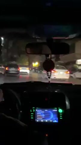 A person is driving a car through city streets at night. The car's dashboard is visible, and a rearview mirror shows the driver. Cars are ahead in traffic, and streetlights illuminate the surroundings. Music is playing in the car.