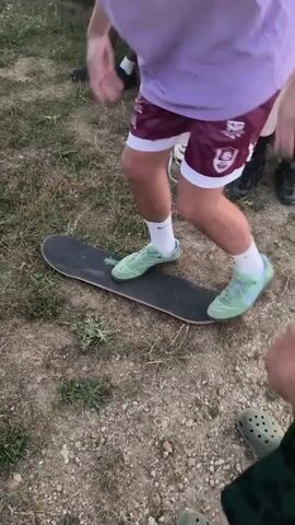 This video shows a group of young people gathered outdoors. The primary subject is a young man wearing a light purple t-shirt, maroon shorts with a white trim and logo, white socks, and light green sneakers. He is positioned on a skateboard, which is lying flat on the ground. The ground appears to be a mixture of dirt, gravel, and dry grass, suggesting an outdoor, possibly unpaved area.

The young man is actively engaged with the skateboard. He is shown in various poses, appearing to be either practicing or demonstrating a skateboarding maneuver. His body is in motion, and his feet are placed on the skateboard. Another person's legs, wearing camouflage Crocs, are visible in the lower right corner of the frame, and another person's legs wearing black shoes and white socks are visible in the background to the left. The presence of other people suggests a social gathering or a casual outing.

The scene appears to be set during daylight, likely afternoon, given the ambient light. The weather seems to be dry. The overall mood seems casual and possibly energetic, indicated by the activity. There is text visible on the logo of the maroon shorts, but it is not clearly legible. The context provided indicates the location as Changis-sur-Marne, France.