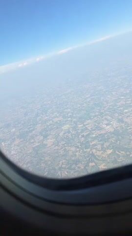 The video shows an aerial view from an airplane window looking out over a landscape below. The sky is a clear blue with a faint haze near the horizon. The landscape below appears to be a patchwork of agricultural fields, with various shades of green and brown indicating different crops or land uses. A small town or city is visible in the distance, with buildings and roads appearing as small details from this altitude. The edge of the airplane window frame is visible in the foreground, providing a sense of perspective. The video is a static shot, with no apparent movement of the camera or subjects.