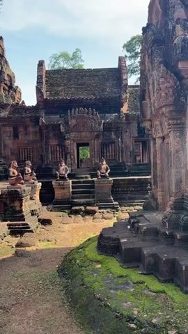The video pans across a courtyard in front of an ancient, weathered stone temple complex. The structures are made of reddish-brown stone, with intricate carvings visible on the walls and towers. Several small, weathered statues of seated figures, possibly guardians or deities, are positioned on raised platforms and along the steps leading to the temple entrance. The ground is uneven, with patches of green moss growing on the stones and soil. Trees and foliage are visible in the background, and the sky above is partly cloudy. The overall impression is one of age and historical significance.
