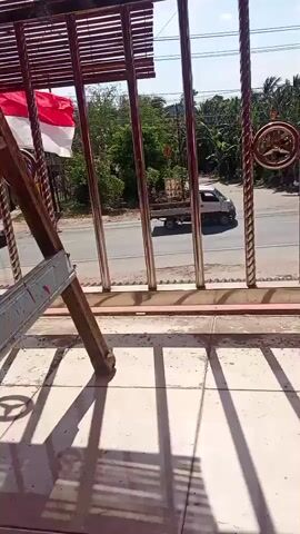 The video pans across a street scene viewed through a metal railing. A red car is visible on the road, and a small white truck is parked further down. Trees and houses line the street, and the sky is bright and cloudy. The camera is positioned on a balcony or porch, with tiled flooring and a shadow pattern cast by the railing. The railing itself is made of shiny metal poles and has a decorative circular element. There is also a glimpse of a red and white flag in the foreground.