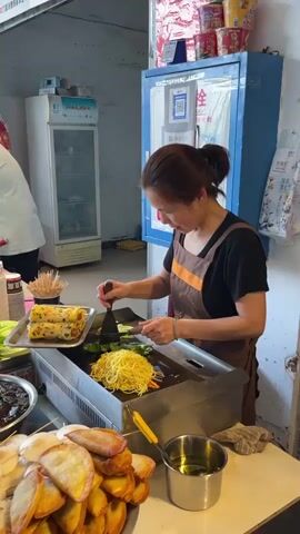 A woman is shown standing behind a street food stall, wearing a brown apron over a dark-colored shirt, her hair pulled back in a ponytail. She is cooking noodles and vegetables on a flat-top grill. The noodles are yellow and the vegetables include green lettuce. She is using a spatula and a small knife to manipulate the food on the grill. To her left is a tray with rolled egg dishes. In front of her is a stack of fried dumplings or savory pastries and a metal container with oil. Behind her are two refrigerators; one is a small white Haier refrigerator, and the other is a taller blue one. The background suggests an outdoor setting near a building.