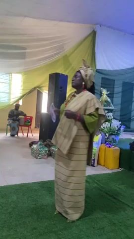 A woman stands on artificial turf in front of a speaker, tables, and what appear to be gifts. She is dressed in a traditional African dress. She is holding what appears to be a small book or pamphlet and gestures with it. The room is decorated with white and green draping and a spotlight illuminates the area behind her.