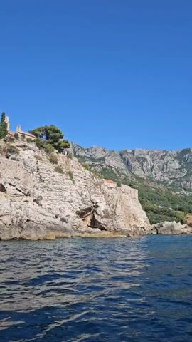 The video shows a boat traveling on the water, with a rocky cliff face on the right side of the frame. There are buildings and trees on top of the cliff. The sky is clear and blue. The water is a deep blue and appears to be slightly choppy. The camera is moving from left to right, showing more of the cliff as it moves.