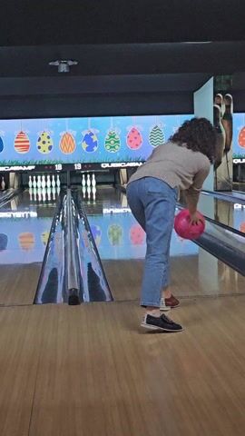 The video shows a woman bowling in a bowling alley. She is facing away from the camera and holds a pink bowling ball. She bends over and releases the ball down the lane.  The ball travels towards the pins, but it's hard to see the result. The bowling alley has a wooden floor and a digital scoreboard with a colorful Easter egg theme. Other lanes are visible in the background. The woman is wearing jeans, a tan sweater, and bowling shoes.