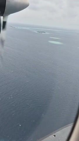 The video shows an aerial view from a plane window, with a propeller in the foreground. The main subject is the vast expanse of the ocean, which has a dark blue, rippled surface under a cloudy sky. Scattered across the ocean are small, light green islands, some with visible structures.