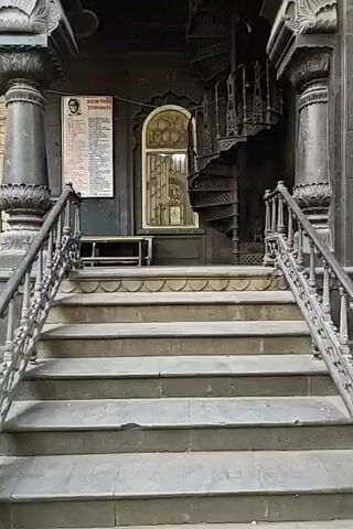 The video showcases a multi-tiered, dark stone building complex with intricate archways and elaborate columns. It begins with a close-up of a staircase leading to a raised platform featuring a spiral staircase on the right. A poster or notice is affixed to the wall on the left. The camera then pans upward, revealing the detailed architecture, including balconies and decorative elements. From a low-angle perspective, the video reveals the structure's height and the presence of arched entrances leading to the outside. People and cars are visible in the background, suggesting the location is accessible to the public. The environment includes trees visible above the building and glimpses of a bright sky.
