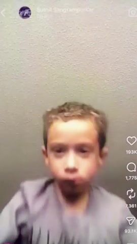 A young boy with curly brown hair is looking into the camera. He is wearing a gray t-shirt with the words "ALWAYS BE" visible. The background is a textured ceiling. The boy opens his mouth wide and then coughs into his fist. He has rosy cheeks and is looking directly at the camera. The video is from a social media platform and displays engagement metrics on the right side of the screen.