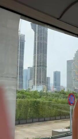 The video shows a view of a city skyline through the window of a moving vehicle, possibly a bus or a train. Several tall buildings are visible, including some under construction and one with a distinctive design featuring a gold tree-like pattern. Greenery is present in the foreground, partially obscuring the view of the buildings. A black lamppost with two lanterns is also visible in the foreground. The sky is overcast, creating a muted and slightly gray atmosphere.  There are no people directly visible in the frame. The camera pans slightly, adjusting the view of the cityscape as the vehicle moves.