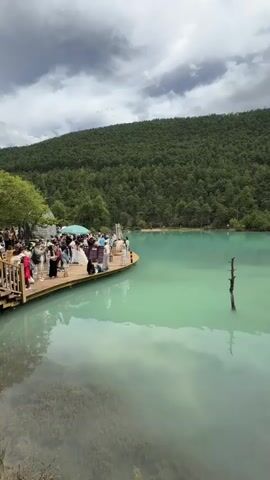 A wide shot shows a teal-colored lake with a person's reflection in the water in the foreground. On the left side of the image, a wooden platform extends into the water, with about a dozen people gathered on it. The people are dressed in casual clothing and appear to be taking photos. In the background, a lush green forest covers a mountain, and the sky is cloudy and gray. The video is shot from a low angle, giving the impression of being on the water's surface.
