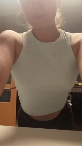 A woman wearing a light blue cropped tank top and black pants is shown from the chest up. She is in what appears to be a kitchen. In the background, a wooden door with glass panels is visible, along with kitchen cabinets and appliances. The lighting is dim, with a bright light source coming from above and to the left. Her hair is pulled back. She is looking down and her mouth is slightly open.