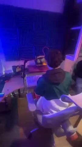 A video shows a recording studio environment with a person sitting in front of a computer. The person has curly hair and is wearing a green and white jacket. The computer is on a desk with a keyboard and microphone. The walls of the room are covered in soundproofing panels. The room is illuminated with blue light. A piece of paper or music sheet can be seen being held in the individual's hands. The camera is at a low angle, showing the back of the person and part of the recording equipment.