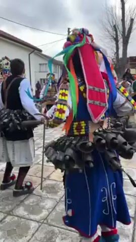 The video shows people dressed in traditional costumes with bells attached to their clothing. The costumes are colorful and elaborate, featuring ribbons and intricate designs. The people are moving, likely dancing or participating in a parade. The environment appears to be a street in a village or small town, with buildings visible in the background. The weather is overcast. The main action is the movement and ringing of the bells as the people in costume move. There is a sense of celebration and tradition evident in the scene.