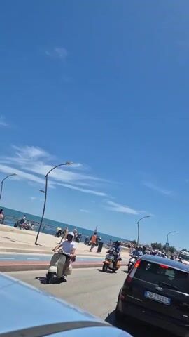 The video shows a street scene with a clear blue sky and some clouds. There is a road with several motorbikes and scooters on it. People are riding the motorbikes and scooters, and some are wearing helmets. There are also some cars visible. In the background, there is a body of water, likely the sea, and a boardwalk or promenade with people walking and sitting. Streetlights are visible along the side of the road. The overall impression is a sunny day in a coastal area with people enjoying a ride on their motorbikes.