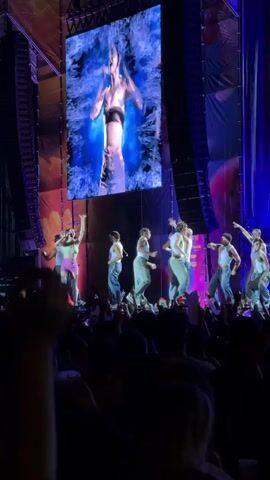 The video shows a live concert performance with a singer on stage, accompanied by several dancers. A large screen behind the performers displays a dynamic visual of a person in water. The stage is illuminated with colorful lights, and a crowd of spectators can be seen in the foreground, many holding up their phones. The overall atmosphere appears energetic and lively, typical of a music event.
