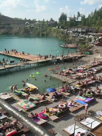 The video shows a clear turquoise lake surrounded by hills and trees under a bright sunny sky with some clouds. A man-made beach area is set up along the edge of the lake with numerous lounge chairs and umbrellas. Many people are visible sunbathing, swimming in the lake, and relaxing on the beach. There are several wooden piers extending into the water, with people standing and sitting on them. Some people are seen swimming and playing in the water, while others are on floating devices. In the distance, there are some boats on the lake. The overall scene depicts a lively outdoor recreational area, likely a popular spot for swimming and leisure activities on a summer day. The camera pans across the scene, capturing various groups of people and activities around the lake and beach.