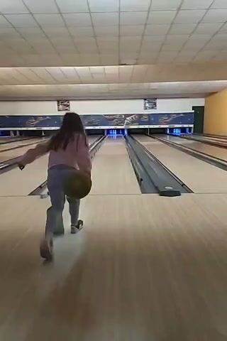 The video shows a young girl bowling in a bowling alley. She is wearing a light pink sweater and blue pants and has long dark hair. She bowls the ball down the lane toward the pins at the end. The alley has multiple lanes and is brightly lit with overhead lighting. The lanes are made of wood and have gutters on either side. Behind the bowling lanes are decorative panels with various designs. The girl follows through with her bowling motion and watches the ball roll down the lane.