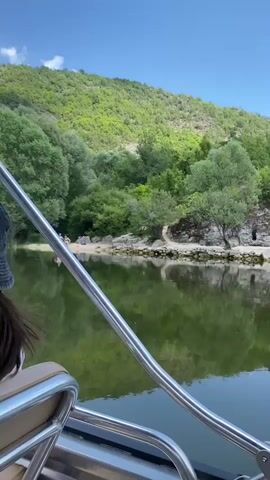 A video taken from a boat shows a calm, green body of water reflecting the surrounding lush green trees and a hill covered in vegetation under a clear blue sky with some clouds. On a sandy shore, three people are visible. Two individuals, a male and a female, are standing and appear to be interacting, possibly posing for a photo as the male holds up a phone. A third person, possibly lying down, is also present on the shore. The boat's metallic railing is prominently featured in the foreground.