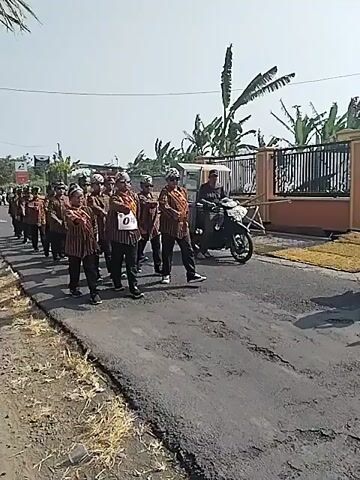 A group of men in traditional striped shirts and head coverings are marching in a procession down a paved road. They appear to be participating in a parade or festival. Some of the men are wearing sashes with numbers on them, suggesting they are part of different groups or teams. A person is riding a motorcycle alongside the procession, possibly providing support or escort. The environment appears to be a rural or suburban area with houses and vegetation visible in the background. The sky is clear and the sun is shining, indicating it is daytime.