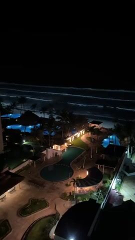 A nighttime aerial view captures a resort complex illuminated beside an ocean. The dark ocean shows visible white lines of breaking waves along a sandy coastline. The resort grounds feature multiple brightly lit blue swimming pools, numerous tall palm trees illuminated from below, green grassy areas, and various resort buildings and pathways lit with warm yellow and white lights. The camera slowly pans from left to right, revealing more of the illuminated resort facilities along the dark beach.
