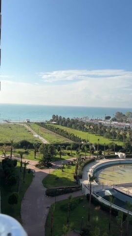 The video shows an elevated view of a park and coastal area. The park consists of green lawns, scattered trees, and paved pathways. In the foreground is a circular concrete structure resembling an empty pool or fountain. The park transitions into a grassy area leading to a beach on the edge of a light blue ocean under a bright blue sky with scattered clouds. A few buildings are visible beyond the park, closer to the coastline. Birds fly in the sky. The overall scene suggests a sunny, calm day.